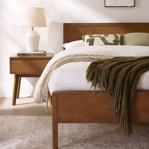 Close-up of cozy minimalist bedroom with mid-century wooden bed frame and nightstand, neutral white bedding, textured green knit throw and patterned accent pillow, ceramic table lamp on wooden bedside table