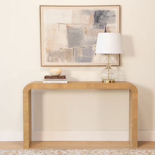 Modern minimalist entryway with a light wood rounded-console table topped by a glass-and-gold table lamp, stacked books, a candle, and neutral abstract framed wall art