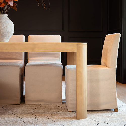 Minimalist modern dining room with light wood dining table, beige upholstered chairs, decorative stone vase and neutral patterned rug against black paneled wall