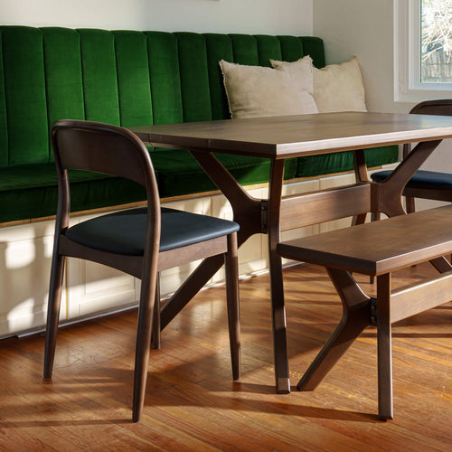 A mid-century modern wooden dining table set with matching chairs and a bench in a bright dining room with a green velvet banquette and wood floors.