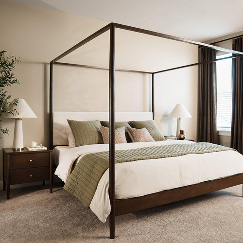 Four-poster canopy bed with dark wooden frame, neutral bedding and green quilted pillows in a modern sunlit bedroom with nightstand, lamp and window curtains
