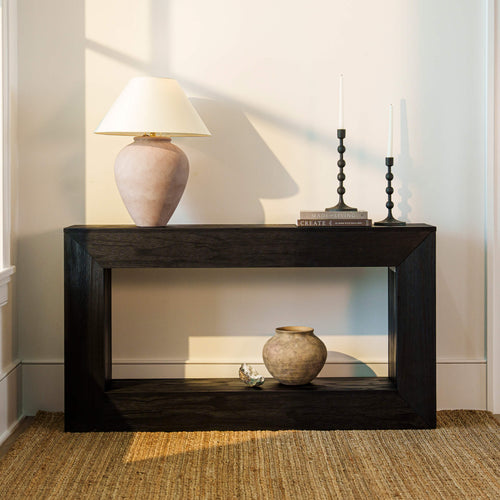 A dark wood console table styled with a beige ceramic table lamp, black candlesticks, a small vase, and a shell, set against a neutral wall with warm sunlight casting shadows.