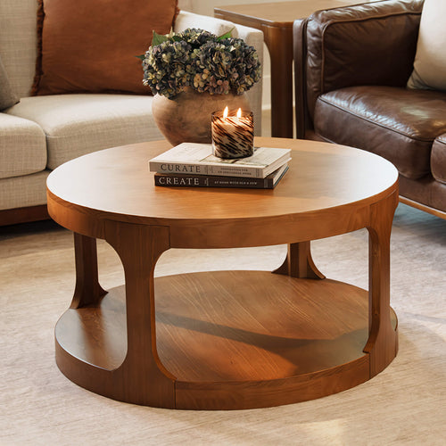 Wooden round coffee table styled with lit patterned candle, stacked home decor books and a rustic vase of hydrangeas beside a neutral fabric sofa and brown leather armchair in a cozy modern living room