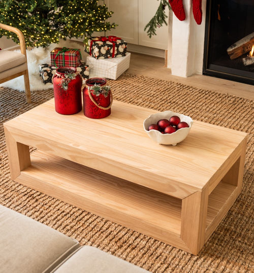 Scandinavian light wood coffee table on jute rug in cozy living room with decorated Christmas tree, wrapped gifts, lit fireplace, red holiday candle jars and ornaments