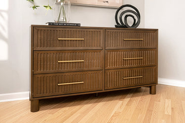 Mid-century modern six-drawer brown wooden dresser with ribbed drawer fronts and brass handles on light hardwood floor in a styled bedroom