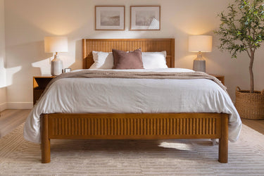 Cozy minimalist bedroom with wooden slatted bed frame, white neutral bedding and beige throw, matching nightstands with lamps, framed art and potted plant