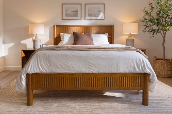 Cozy minimalist bedroom with wooden slatted bed frame, white neutral bedding and beige throw, matching nightstands with lamps, framed art and potted plant