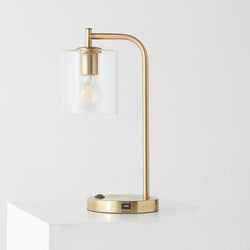 Modern brass desk lamp with clear glass shade, exposed filament Edison bulb and USB charging port on a minimalist white pedestal