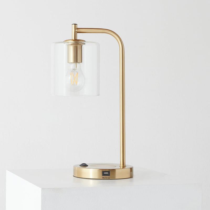 Modern brass desk lamp with clear glass shade, exposed filament Edison bulb and USB charging port on a minimalist white pedestal