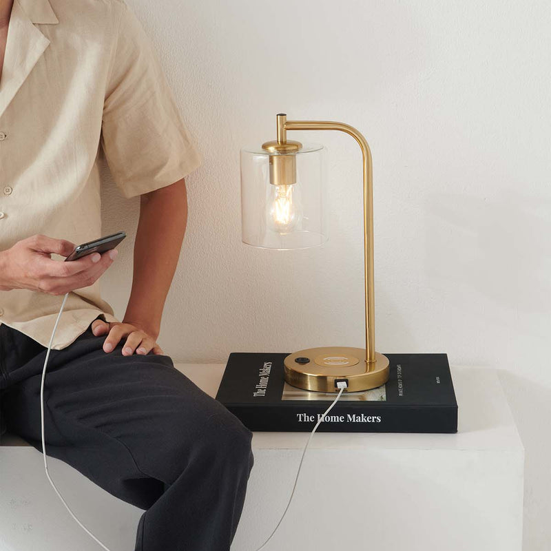 Minimalist bedside: brass table lamp with clear glass shade and Edison bulb on a book, smartphone USB charging.