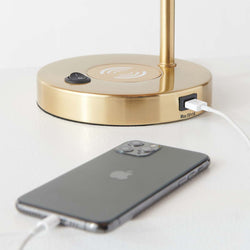 Modern gold desk lamp with built-in wireless charging pad and USB port, smartphone charging on white bedside table