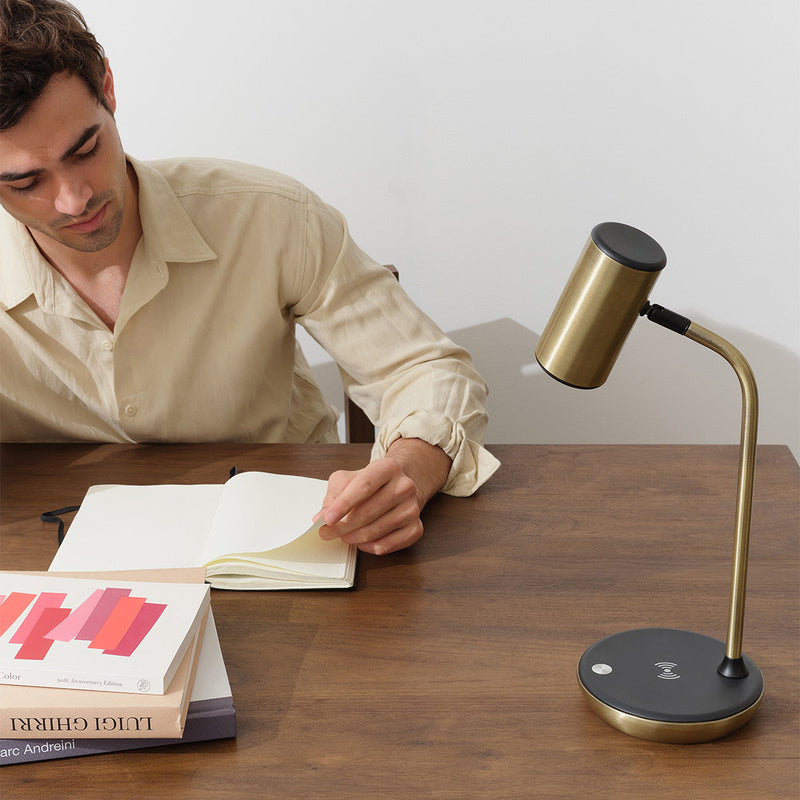 Man studying at a wooden desk, turning pages in a notebook beside a modern brass desk lamp with wireless charging base and stacked design books in a minimal home office workspace