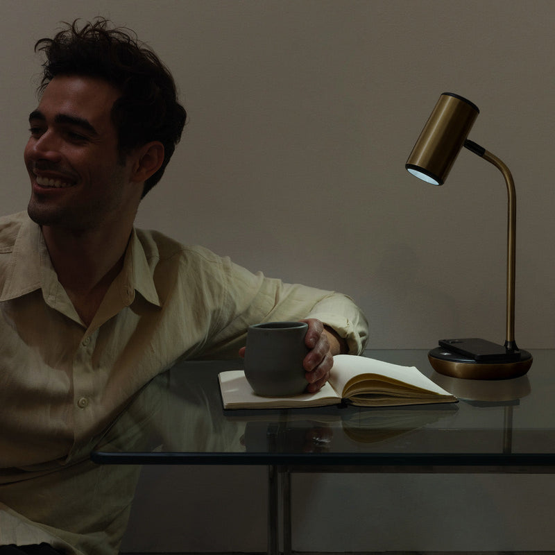 Smiling man sitting at a glass desk holding a coffee mug with an open notebook under a modern brass desk lamp in a cozy home office ambiance