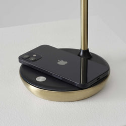 Black iPhone on brass-accented wireless charging stand on white textured table, minimalist desk accessory