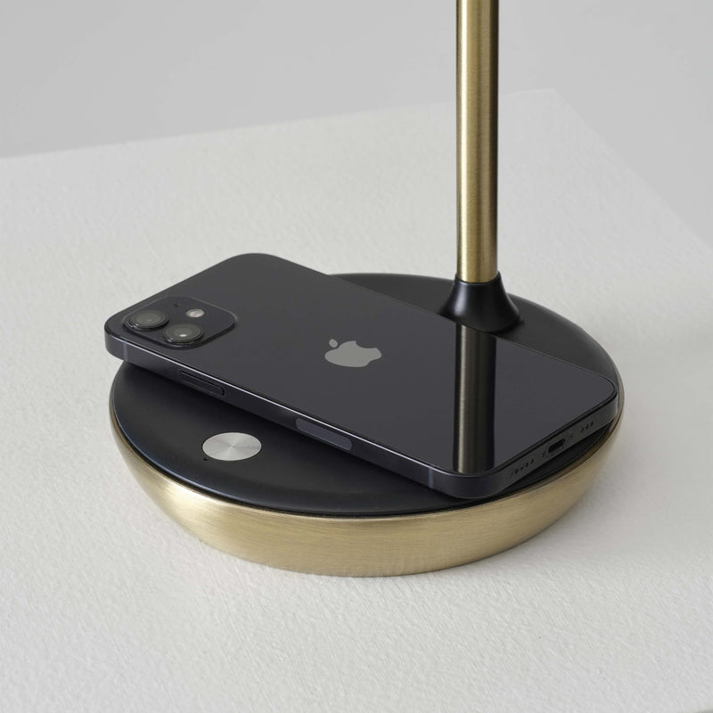 Black iPhone on brass-accented wireless charging stand on white textured table, minimalist desk accessory