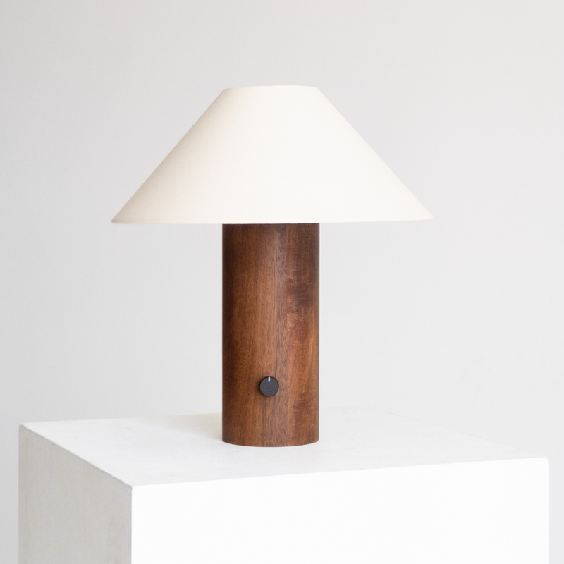 Minimalist modern table lamp with cylindrical walnut wood base and white conical fabric shade on a white pedestal