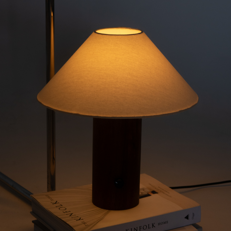 Modern wooden-base bedside table lamp with beige fabric shade casting warm ambient light on stacked Kinfolk books