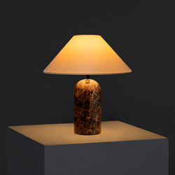 Modern marble table lamp with beige fabric shade casting warm ambient light on a white pedestal against a dark background
