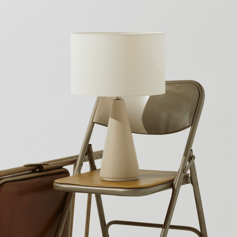 Metal folding chair with contemporary beige table lamp and suitcase against neutral background.