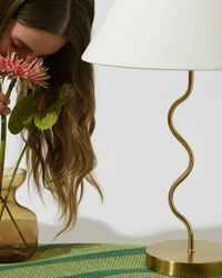Woman smelling pink flowers near a gold lamp on a green woven tablecloth.