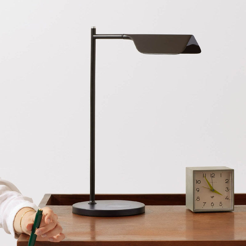 Modern black desk lamp on wooden table with minimalistic clock and person holding a pen.