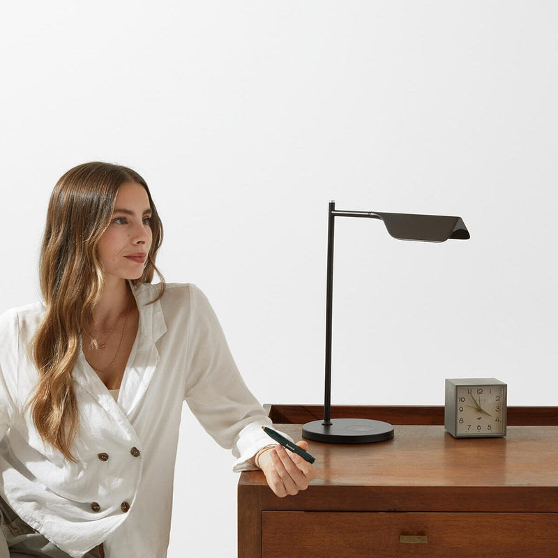 Woman in white blouse sitting at a wooden desk with modern black desk lamp and clock.