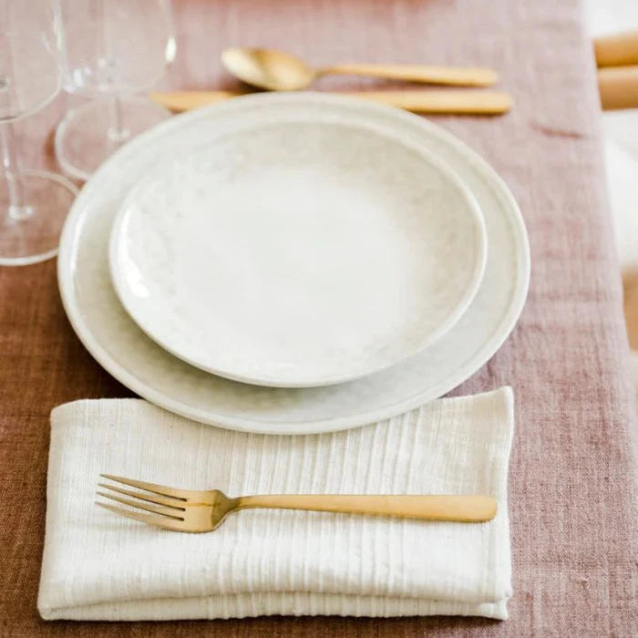 Elegant dinner table setting with white ceramic plates, gold cutlery, and a folded napkin on a textured tablecloth.