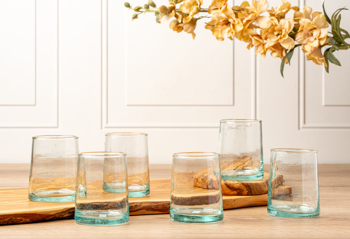 Six clear glass tumblers on a wooden serving tray with beige flowers in the background.