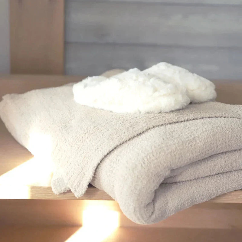 Cozy beige bath towel with fluffy white slippers on a wooden shelf, sunlight streaming in.