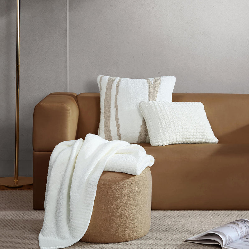 Cozy modern living room with beige couch, textured white pillows, soft throw blanket, and carpeted floor.