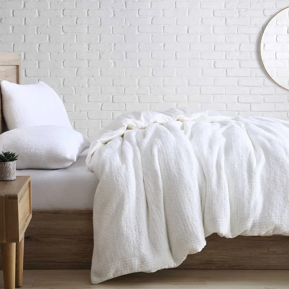 Luxurious white bedding set on a wooden bed frame in a minimalist bedroom with a white brick wall background.