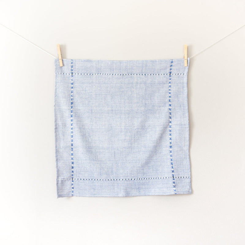 Light blue cotton linen napkin with decorative stitching hanging on a clothesline against a white background.
