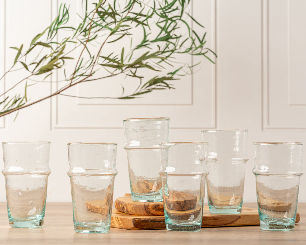 Eight eco-friendly glass tumblers with textured design on a wooden surface, in front of a white paneled wall with green foliage.