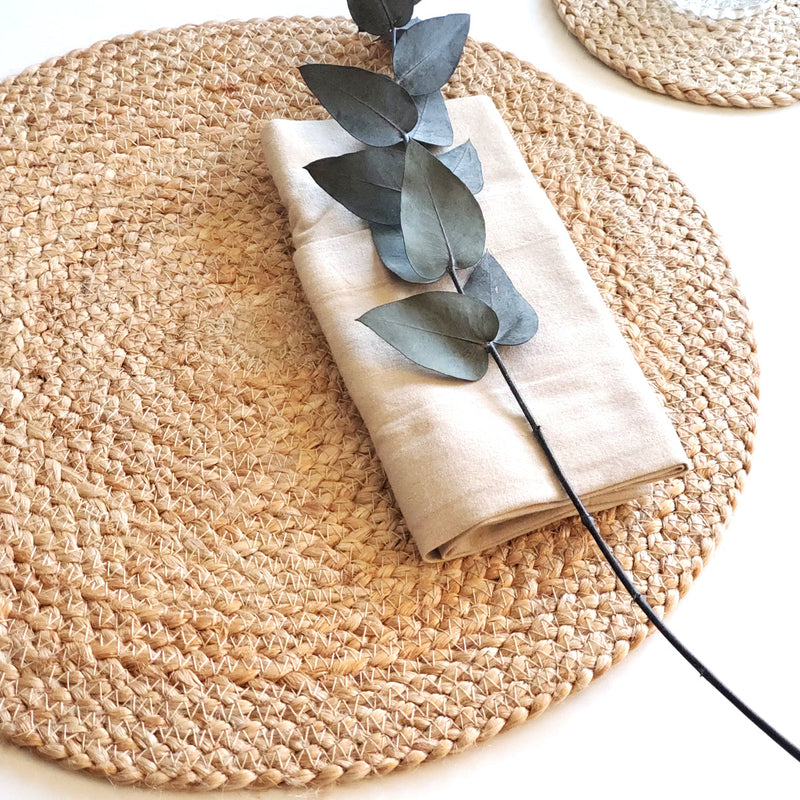 Woven placemat on a table with a folded beige napkin and eucalyptus branch for rustic dining decor.