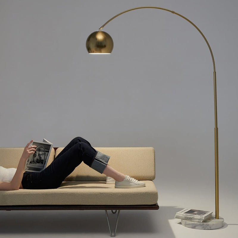 Person relaxing on a modern sofa under a stylish brass floor lamp, reading a book in a minimalist setting.