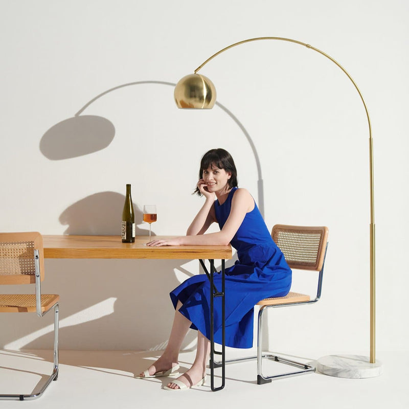 Woman in blue dress sitting at modern dining table with wine, under stylish gold floor lamp.