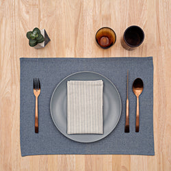 Neatly arranged table setting with blue placemat, gray plate, striped napkin, black cutlery, and brown glasses on a wooden table surface.