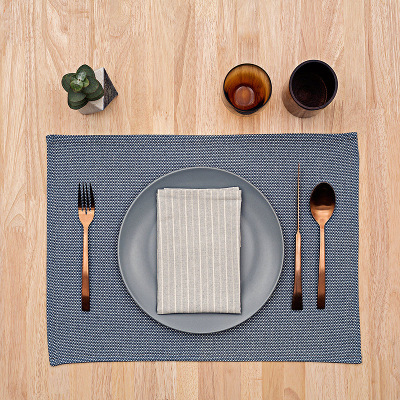 Neatly arranged table setting with blue placemat, gray plate, striped napkin, black cutlery, and brown glasses on a wooden table surface.