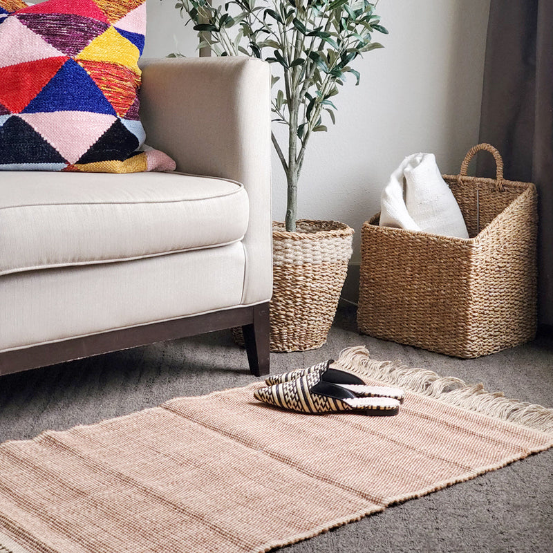 Cozy living room interior with beige armchair, colorful geometric pillow, woven basket, decorative green plant, and soft area rug.