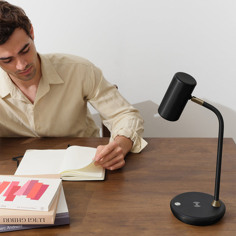 Man writing in a notebook beside a modern black desk lamp on a wooden table.