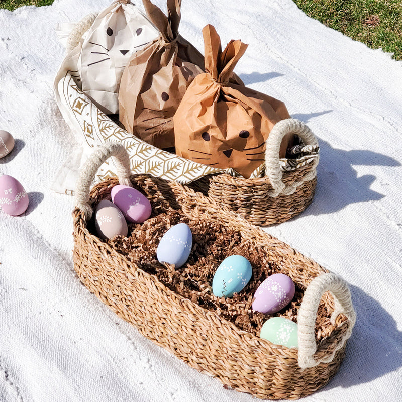 Woven baskets with decorated Easter eggs and bunny-themed gift bags on a white picnic blanket in a garden setting.