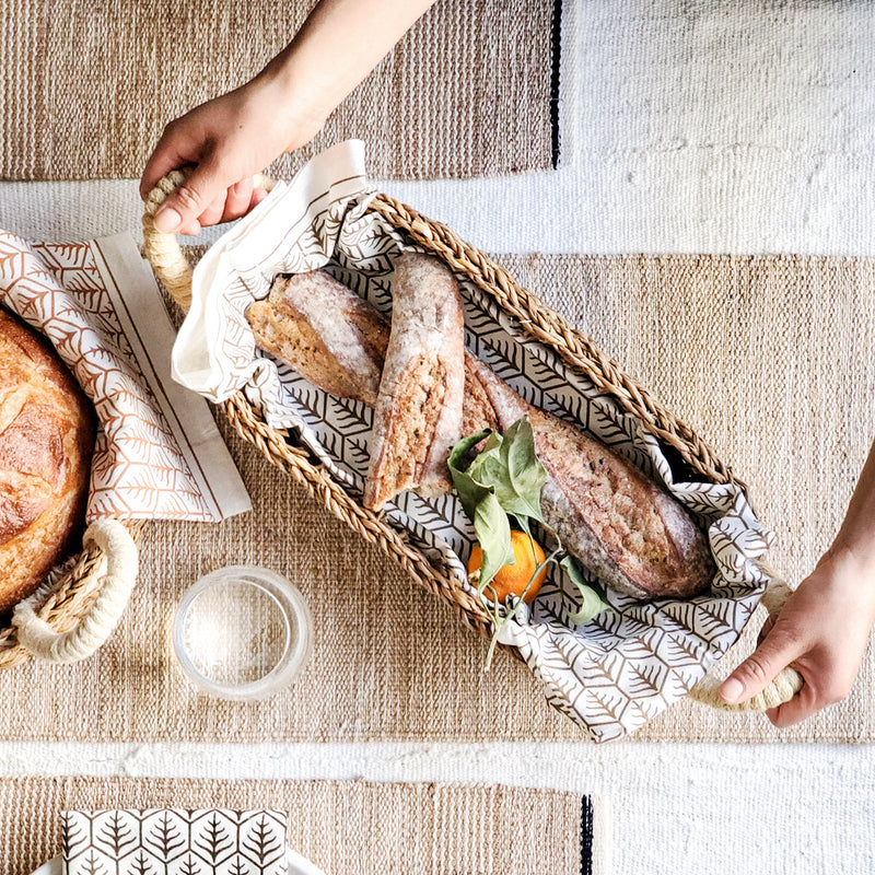 Hand holding a woven basket with fresh baguettes and a citrus fruit, perfect for a rustic dining table setting.
