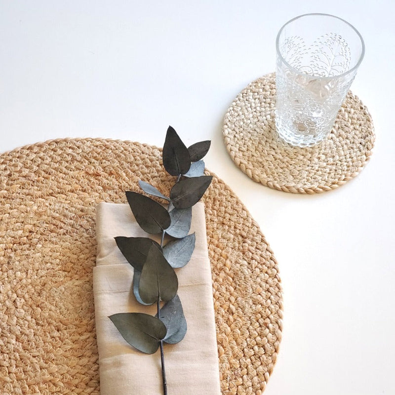 Woven placemat with a folded beige napkin and eucalyptus branch, next to a textured glass on a matching coaster.