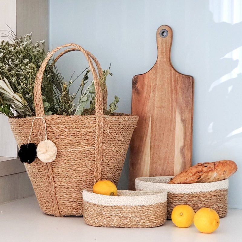 Handwoven jute basket with dried flowers and lemons next to wooden cutting board and bread, kitchen decor.