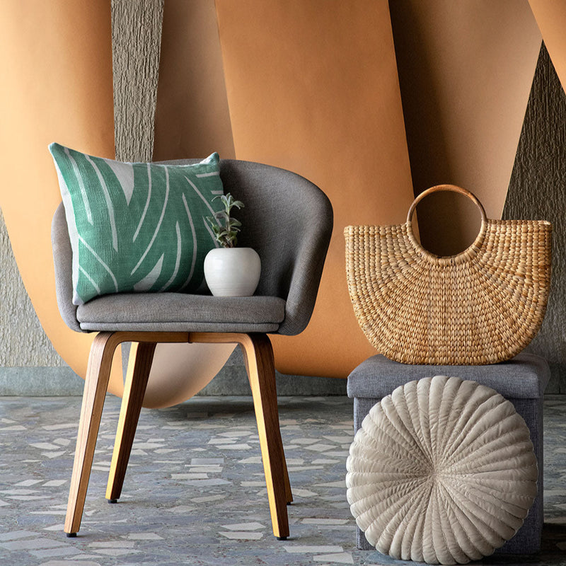 Modern interior design with a stylish gray chair, green botanical cushion, woven handbag, and textured cushion on a patterned floor.