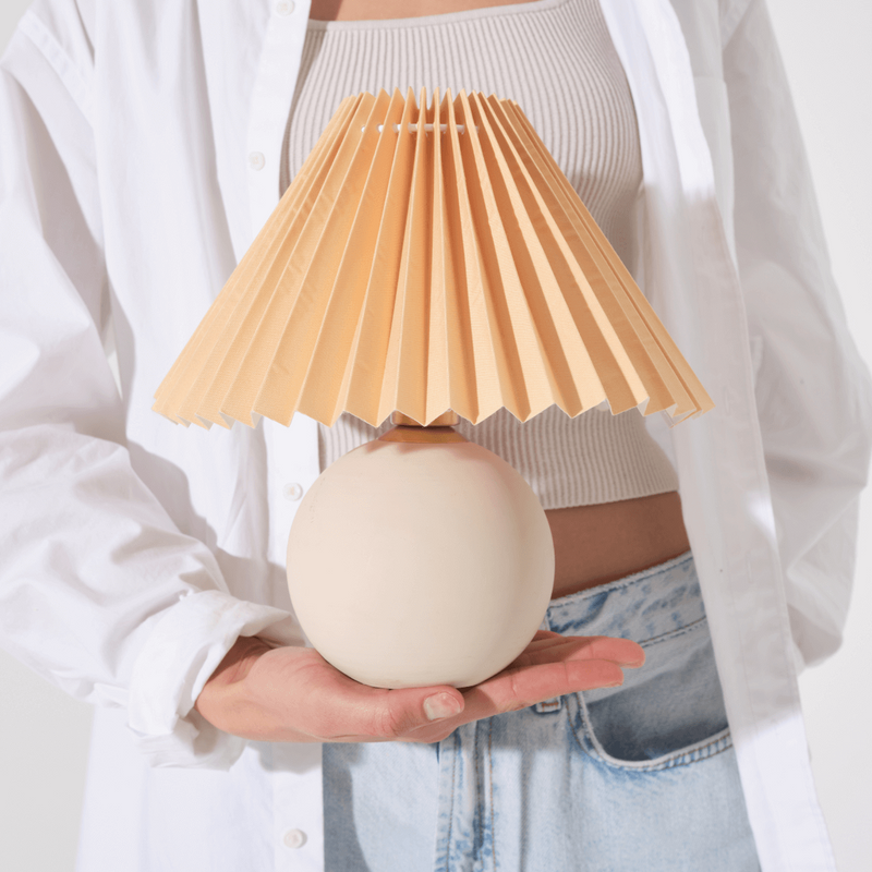Person holding a modern table lamp with a beige pleated shade and round ceramic base.