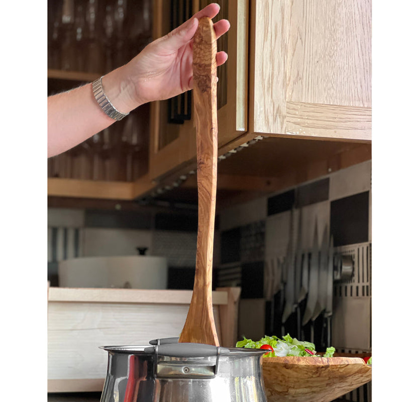 Hand holding olive wood spatula above a stainless steel pot in a modern kitchen setting.