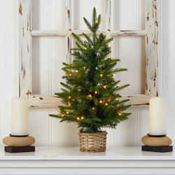 Small tabletop Christmas tree with warm white string lights in a wicker basket on a white mantel, rustic distressed window frame backdrop and two pillar candles, holiday decor
