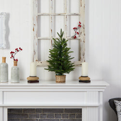 Small potted Christmas tree in a wicker basket on a rustic white mantel with pillar candles, ceramic vases, red berry accents and distressed window frame