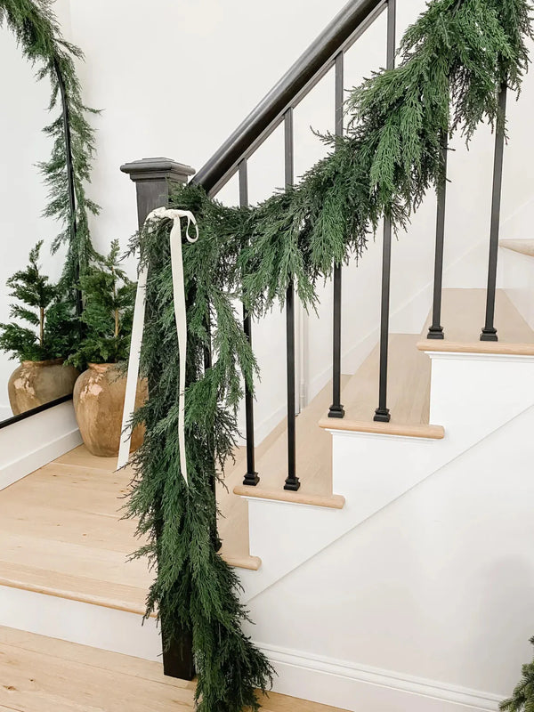 Christmas staircase garland draped on black iron railing with white ribbon, lush cedar greenery and light oak steps in a neutral modern farmhouse entryway